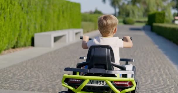 おもちゃの電気自動車に乗って日当たりの良い公園の道を眺める子供のバックビュー 高品質の4K映像 — ストック動画