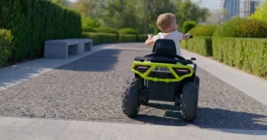 Güneşli bir park yolunda oyuncak elektrikli bir arabayla gezen küçük bir çocuğun arka görüntüsü. Yüksek kalite 4k görüntü