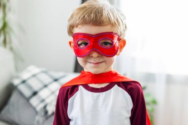 Pelerinli ve maskeli, kendinden emin bir şekilde gülümseyen bir süper kahraman gibi giyinmiş genç çocuk..