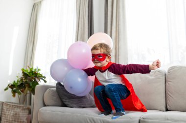 Kırmızı pelerinli ve maskeli süper kahraman kostümlü çocuk balonlarla oynuyor. Ev içi boş zaman aktivitesi. Çocukluk hayal gücü ve tasarım ve baskı için oyun konsepti