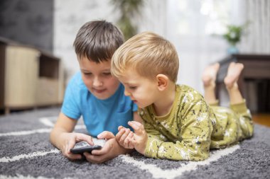 Halıda uzanmış akıllı telefon kullanan iki genç çocuk. Kapalı alan eğlencesi, dijital çocukluk ve tasarım ve baskı kardeşliği konsepti