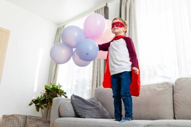 Kırmızı pelerinli ve maskeli süper kahraman kostümlü çocuk balonları tutuyor. Ev portresi gibi. Çocukluk hayal gücü ve oyun konsepti. Tebrik kartı, poster ve davetiye tasarımı