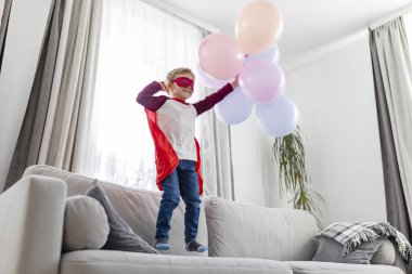 Süper kahraman gibi giyinmiş genç bir çocuk gururla kanepede duruyor, renkli balonlarla oynuyor..