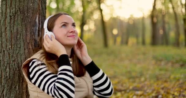 Молода Жінка Насолоджується Прослуховуванням Музики Білими Навушниками Нахиляючись Навпроти Дерева — стокове відео
