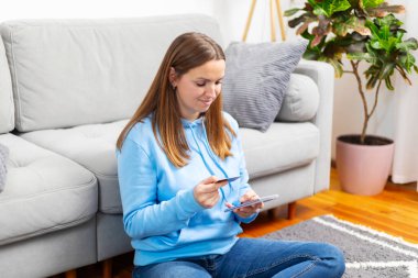 Yerde oturan kadın rahat bir oturma odasında online alışveriş için cep telefonu ve kredi kartı kullanıyor. E-ticaret ve ev alışverişi konsepti. Pankart, web sayfası başlığı veya el ilanı için tasarım.