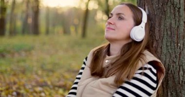 Ağaca yaslanmış beyaz kulaklıkla müzik dinleyen bir kadın. Müzik ve eğlence konsepti. Yüksek kalite 4k görüntü
