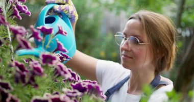 Bardaklı ve önlüklü bir kadın bahçedeki mavi sulama kutusuyla canlı mor çiçekleri suluyor..