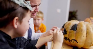 Kostümlü bir çocuk annesiyle balkabağı boyuyor. Cadılar Bayramı el işi ve aile bağları. Yüksek kalite 4k görüntü