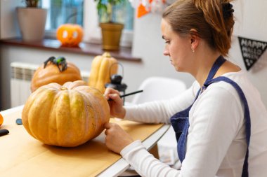 Kadın bir masada büyük bir balkabağı oyuyor, arka planda diğer süslü balkabaklarıyla Cadılar Bayramı şenlikleri için hazırlanıyor..