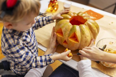 Çocuk, Cadılar Bayramı süslemeleriyle çevrili bir masada, üçgen gözlü büyük bir balkabağını oymaya yardım ediyor. Cadılar Bayramı boyunca kapalı aile aktiviteleri.