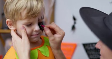 Cadı kostümü giymiş bir kadın Cadılar Bayramı için genç bir oğlanın yüzünü boyuyor ve eğlenceli ve şenlikli bir ortam yaratıyor. Yüksek kalite 4k görüntü