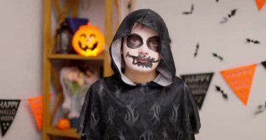 İskelet makyajlı ve pelerinli genç bir çocuk arka planda festival dekorasyonlarıyla ürkütücü bir Cadılar Bayramı kutlamasında ellerini kaldırıyor. Yüksek kalite 4k görüntü