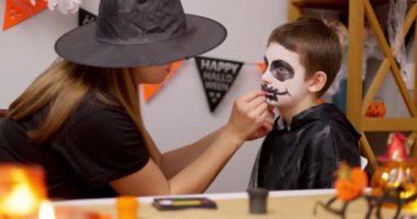 Cadı şapkalı bir anne Cadılar Bayramı kutlaması için genç oğullarının yüzünü siyah beyaz makyajla dikkatlice boyar. Yüksek kalite 4k görüntü