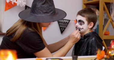 Cadı şapkalı bir anne Cadılar Bayramı kutlaması için genç oğullarının yüzünü siyah beyaz makyajla dikkatlice boyar. Yüksek kalite 4k görüntü