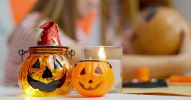 Cadılar Bayramı temalı cam kavanoz, üzerinde balkabağı suratlı ve masada mum var. Arka plana balkabağı çizen bulanık bir kadınla yakın çekim. Cadılar Bayramı kutlaması ve dekorasyon konsepti