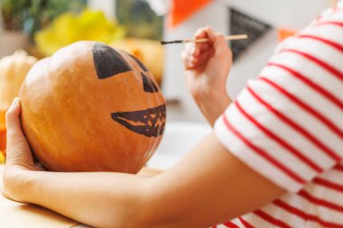 Turuncu bir balkabağına korkunç bir yüz çizen ellerin yakın çekimi. Seçici odaklı yakın plan fotoğraf. Cadılar Bayramı hazırlığı konsepti. Davet için tasarım, poster.