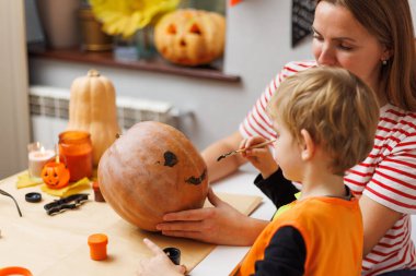Anne ve küçük çocuk süslü bir cadılar bayramı el sanatları masasında balkabağı boyuyor. Aile bağı ve Cadılar Bayramı tasarım ve selamlama hazırlığı konsepti.