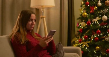 Kırmızı kazaklı genç bir kadın şık bir odada süslü bir Noel ağacının yanında otururken akıllı telefonunu kullanıyor. Yüksek kalite 4k görüntü