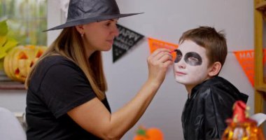 Bir kadın temalı bir odada genç bir çocuğa Cadılar Bayramı makyajı yapıyor ve yüzünü siyah maske tasarımı ile süslüyor. Yüksek kalite 4k görüntü
