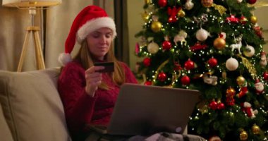 Noel Baba şapkalı bir kadın online Noel alışverişi için dizüstü bilgisayar ve kredi kartı kullanıyor, süslü bir Noel ağacının yanında oturuyor. Yüksek kalite 4k görüntü