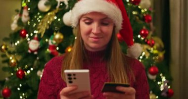 Elinde akıllı telefon ve kredi kartı olan Noel Baba şapkalı bir kadın süslü bir Noel ağacının yanında internetten alışveriş yapıyor. Yüksek kalite 4k görüntü