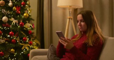 Kırmızı kazaklı genç bir kadın şık bir odada süslü bir Noel ağacının yanında otururken akıllı telefonunu kullanıyor. Yüksek kalite 4k görüntü
