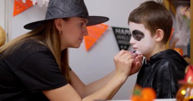 Cadı şapkalı bir anne Cadılar Bayramı kutlaması için genç oğullarının yüzünü siyah beyaz makyajla dikkatlice boyar. Yüksek kalite 4k görüntü
