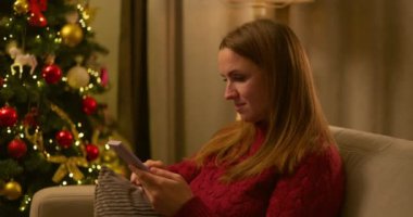 Kırmızı kazaklı genç bir kadın parlak dekore edilmiş bir Noel ağacının yanında otururken akıllı telefonunu karıştırırken gülümsüyor. Yüksek kalite 4k görüntü