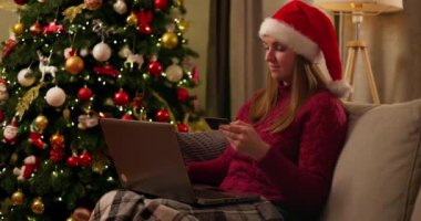 Noel Baba şapkası takan gülümseyen kadın Noel ağacının yanındaki koltukta dizüstü bilgisayar ve kredi kartıyla alışveriş yapıyor. Tatil sezonu ve Noel alışverişi konsepti. Yüksek kalite 4k görüntü