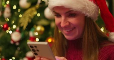 Noel Baba şapkası takan ve akıllı telefonuna süslü bir Noel ağacının önünde kırmızı süveter giyen bir kadın. Yüksek kalite 4k görüntü