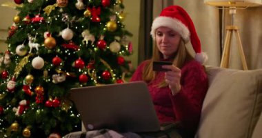 Noel Baba şapkalı bir kadın online Noel alışverişi için dizüstü bilgisayar ve kredi kartı kullanıyor, süslü bir Noel ağacının yanında oturuyor. Yüksek kalite 4k görüntü
