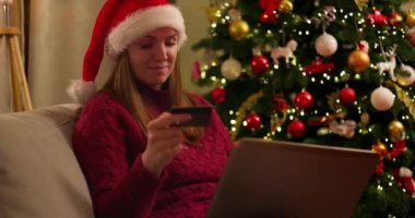 Noel Baba şapkası takan gülümseyen kadın Noel ağacının yanındaki koltukta dizüstü bilgisayar ve kredi kartıyla alışveriş yapıyor. Tatil sezonu ve Noel alışverişi konsepti. Yüksek kalite 4k görüntü