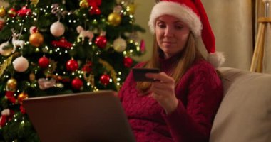 Noel Baba şapkalı bir kadın online Noel alışverişi için dizüstü bilgisayar ve kredi kartı kullanıyor, süslü bir Noel ağacının yanında oturuyor. Yüksek kalite 4k görüntü