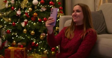 Gülümseyen bir kadın, parlak ışıklarla süslenmiş bir Noel ağacının yanında oturmuş akıllı telefonuyla sohbet ediyor. Yüksek kalite 4k görüntü