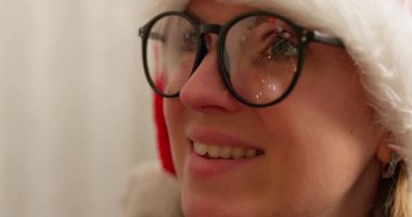 Gözlük takan ve Noel Baba şapkalı bir kadının yakın plan görüntüsü, lenslerine yansıyan Noel ışıkları, neşeli bir atmosfer yaratıyor. Yüksek kalite 4k görüntü
