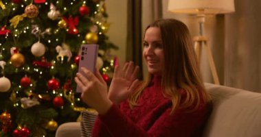 Kırmızı kazaklı bir kadın, arka planında güzel süslenmiş bir Noel ağacı olan neşeli bir video görüşmesinden hoşlanıyor. Yüksek kalite 4k görüntü