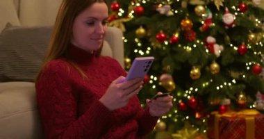 Elinde akıllı telefon ve kredi kartı olan bir kadın süslü Noel ağacının yanında ışık ve süslemelerle oturuyor. Tatil sezonunda online alışveriş. Kapalı mekan sahnesi. Yüksek kalite 4k görüntü