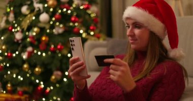 Noel Baba şapkası takan bir kadın süslü Noel ağacının önünde akıllı telefon ve kredi kartı kullanıyor. Tatil alışverişi kavramı. Yüksek kalite 4k görüntü