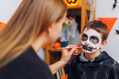 Cadılar Bayramı kutlaması için bir erkeğin yüzüne iskelet yüz boyası süren bir kadın. Çocuk arkasında Cadılar Bayramı süslemeleri olan koyu renk bir kostüm giyiyor.