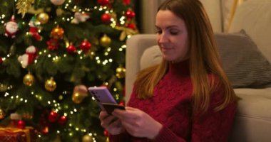 Kırmızı kazaklı bir kadın internetten alışveriş yapmak için akıllı telefon ve kredi kartı kullanıyor, süslü bir Noel ağacının yanında oturuyor. Yüksek kalite 4k görüntü