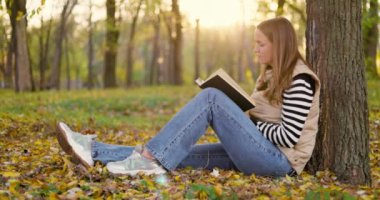 Genç bir kadın, huzurlu bir parkta canlı sonbahar yapraklarıyla çevrili bir kitaba dalmış, bir ağaca yaslanmış, sakin bir şekilde oturuyor. Yüksek kalite 4k görüntü