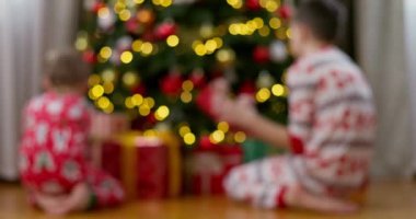 Şenlikli pijamalı iki çocuk süslerle ve paketlenmiş hediyelerle süslü bir Noel ağacının altında hediyeler açıyor. Tatil kutlaması ve aile geleneği kavramı. Yüksek kalite 4k görüntü