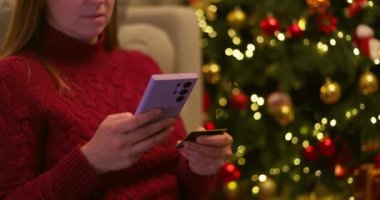 Kırmızı kazaklı bir kadın akıllı telefon ve kredi kartı tutuyor ve ışıkları olan bir Noel ağacının yanında online alışveriş yapıyor. Yüksek kalite 4k görüntü