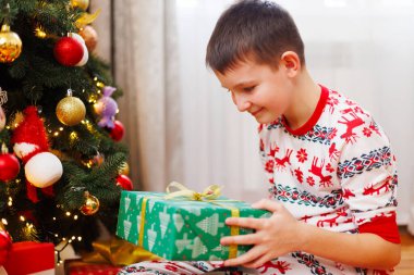 Şen şakrak pijama giyen, Noel ağacının yanında oturan ve altın kurdeleli yeşil bir hediye kutusu açan bir çocuk. Arka planda süslerle süslenmiş Noel ağacı