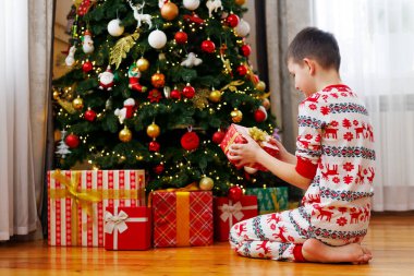Noel ağacının altında diz çökmüş, elinde hediye paketiyle ağacın etrafına yerleştirilmiş, neşeli pijamalı genç bir çocuk. Tasarım için Noel sabahı konsepti