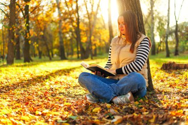 Sonbahar boyunca parkta yerde oturan bir kadın kitap okurken bir ağaca yaslanıyor. Arkaplanda ağaçların arasından süzülen sarı yapraklar ve güneş ışığı yer alıyor.
