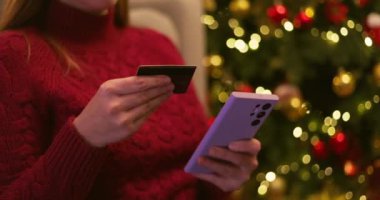 Kırmızı kazaklı bir kadın akıllı telefon ve kredi kartı tutuyor ve ışıkları olan bir Noel ağacının yanında online alışveriş yapıyor. Yüksek kalite 4k görüntü