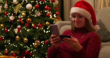Noel Baba şapkası takan bir kadın süslü Noel ağacının önünde akıllı telefon ve kredi kartı kullanıyor. Tatil alışverişi kavramı. Yüksek kalite 4k görüntü