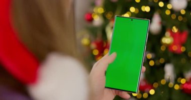 Süslü, bulanık Noel ağacının önünde elinde yeşil ekranlı akıllı telefon tutan bir kadın. Model ve tasarım için yakın çekim. Yüksek kalite 4k görüntü