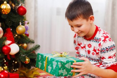 Süslü bir Noel ağacının yanında otururken elinde fiyonklu bir Noel hediyesi tutan neşeli pijamalı gülen çocuk. Tatil döneminde ağaç ışıklarıyla kapalı mekan sahnesi.